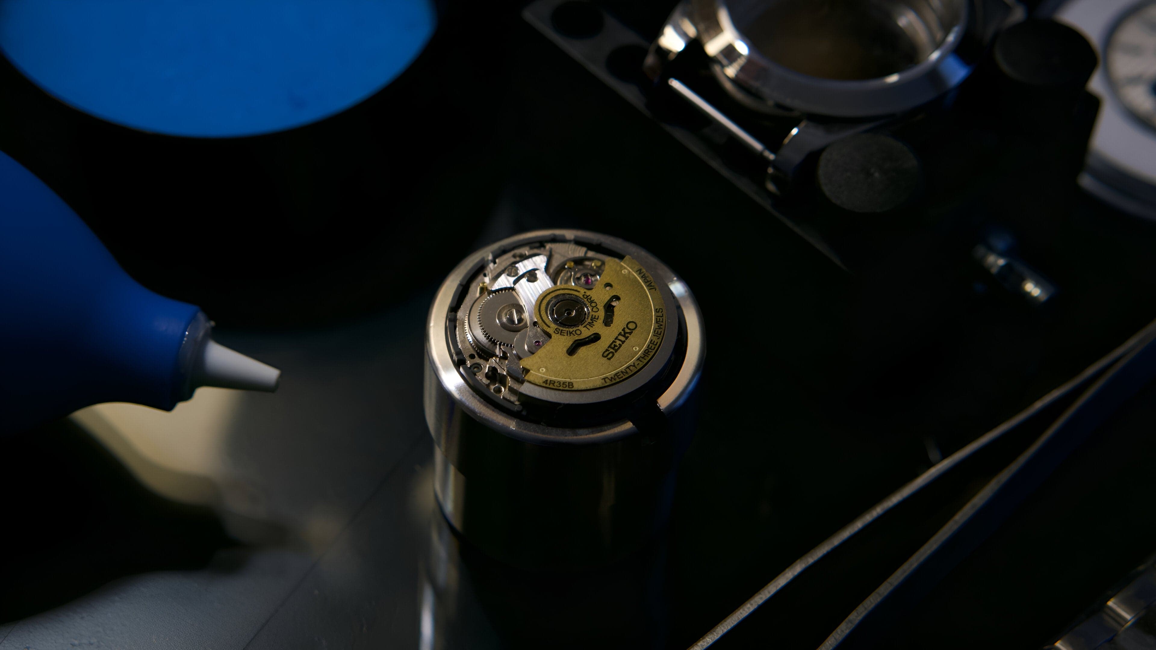 A movement held in a case holder on the bench, a blower and loupe beside it